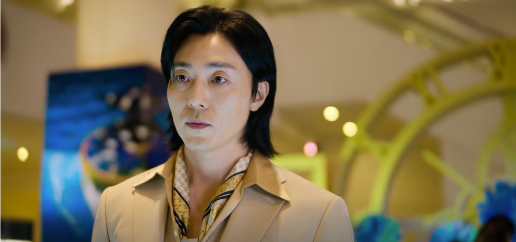 Lee Moo-saeng in a beige suit jacket and patterned scarf in a scene from As You Stood By